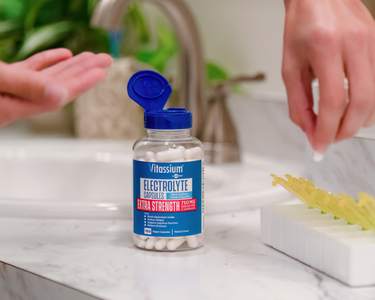 Vitassium Electrolyte Capsules Extra Strength with 750 mg sodium, open bottle on a bathroom counter as a person pours capsules into their hand.