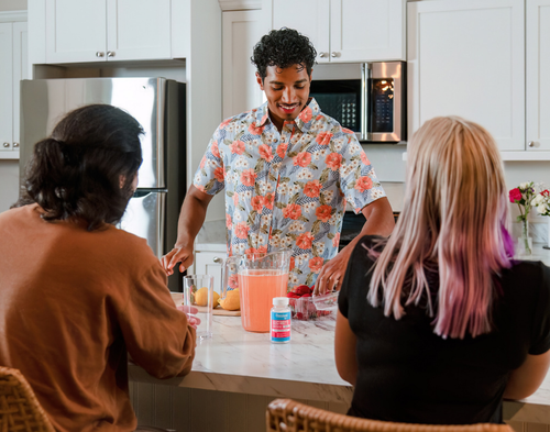 Man in a kitchen making a pitcher of strawberry lemonade with fresh strawberries and lemons, with a bottle of Vitassium Strawberry Lemonade Electrolyte FastChews on the counter in front of the pitcher as two friends watch.