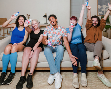 Five diverse people sitting on a couch cheering and holding Vitassium electrolyte capsules and FastChews bottles, with one woman wearing blue compression socks.