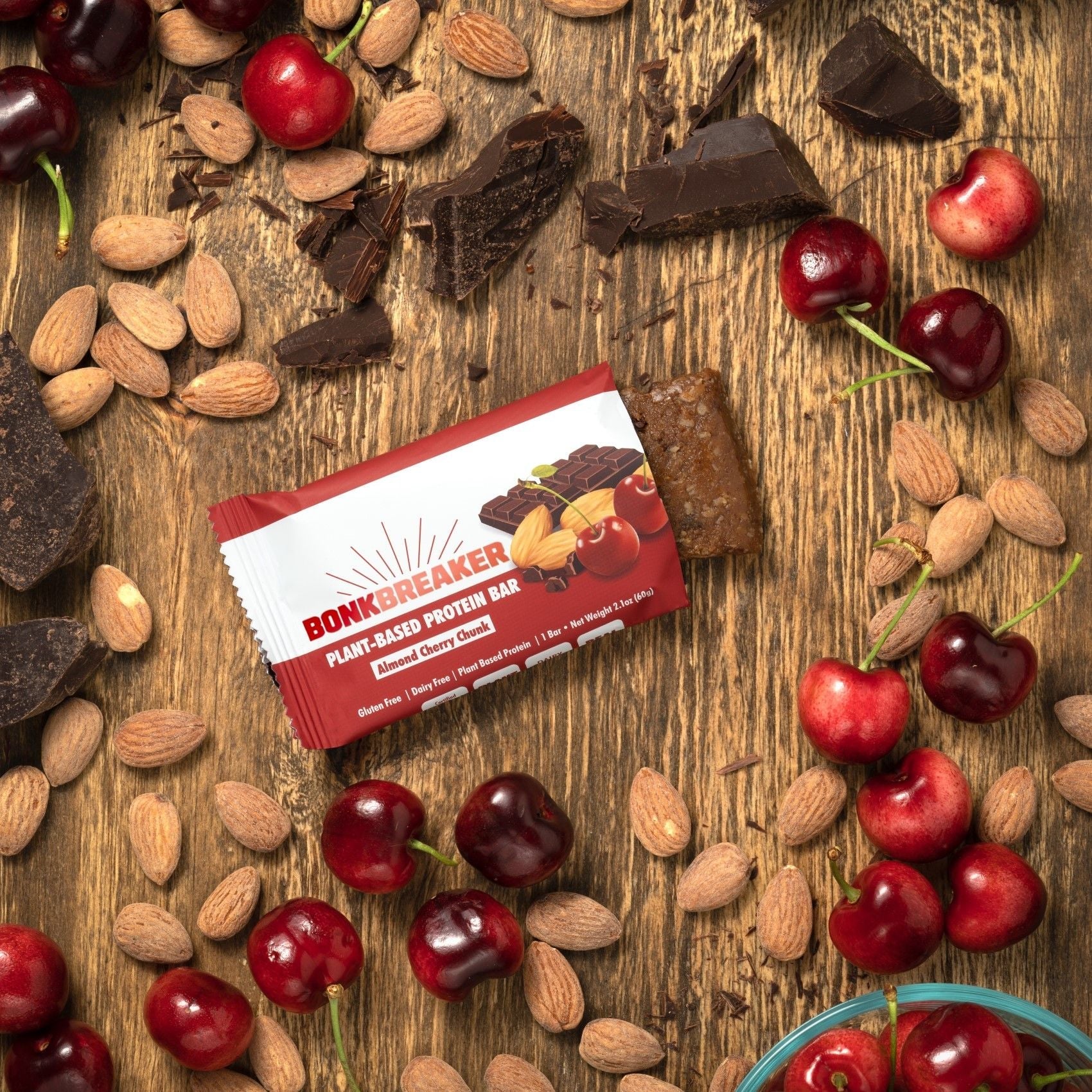Bonk Breaker Almond Cherry Chunk Plant-Based Protein Bar on a table with almonds, cherries, and chocolate